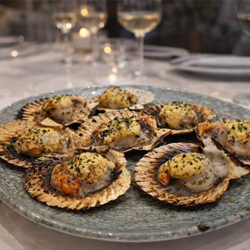 Zamburiñas a la brasa con mantequilla de hierbas servidas en mesa de restaurante con copas de vino desenfocadas, cocina tradicional de producto en Tolón Tolón Bilbao.
